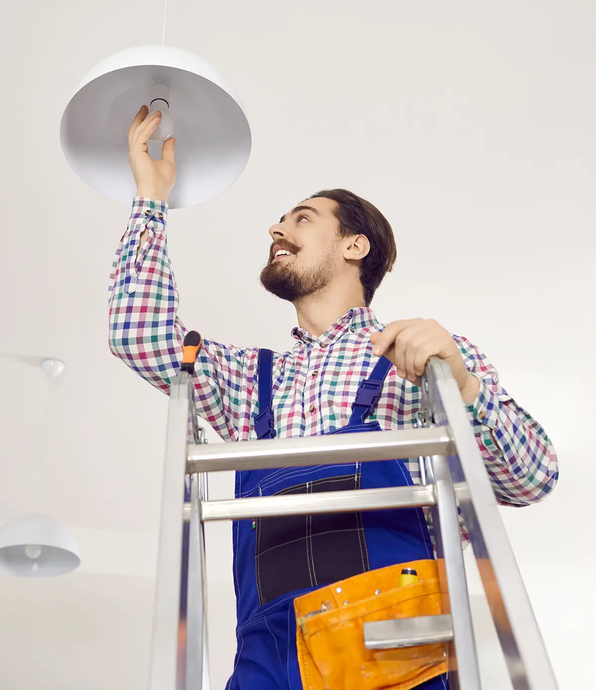 Hausmeister schraubt zu Hause eine Glühbirne ein. Glücklicher Mann in Arbeitskleidung steht auf einer Leiter in einem Wohnhaus, Büro, einer Schule oder einem Krankenhaus und wechselt eine LED-Glühbirne in einer modernen weißen Lampe an der Decke aus.